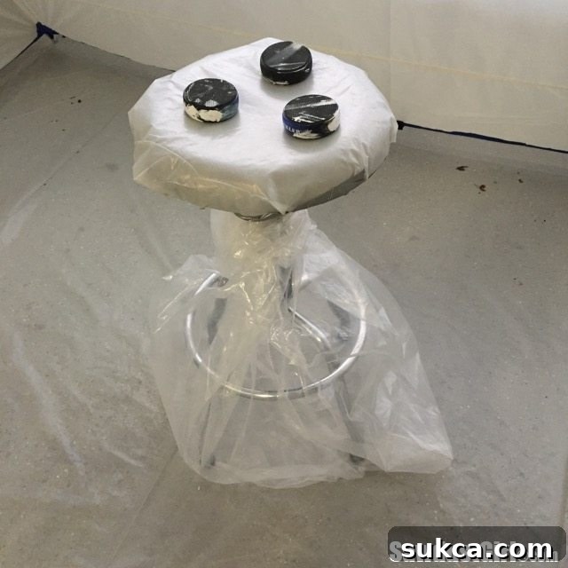 A DIY swiveling paint stand created from a shop stool, wrapped in plastic and using work grippers to hold cabinet doors securely for spray painting.
