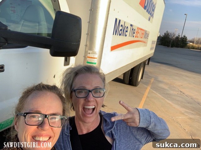 Two women smiling and posing in front of a large moving truck, ready for their cross-country adventure.